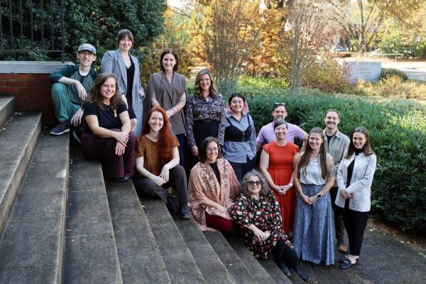 Several people in professional dress standing and kneeling on the front steps of a building with fall foliage in the background.